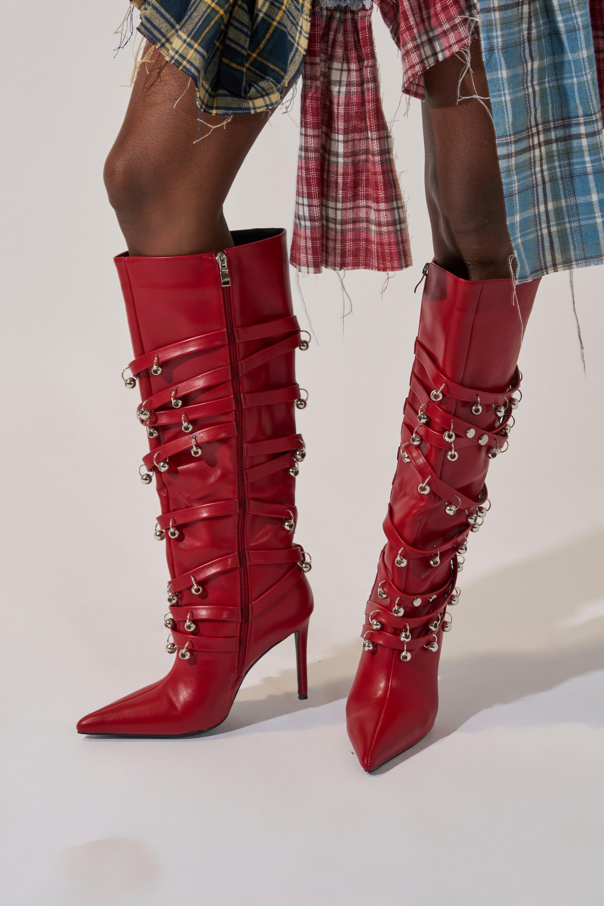 A person wears the AZALEA WANG TIMPANI RED WESTERN FRINGE STILETTO BOOT with a plaid patchwork skirt featuring frayed edges. The bold, knee-high boots with fringe and silver accents complete the eclectic look.
