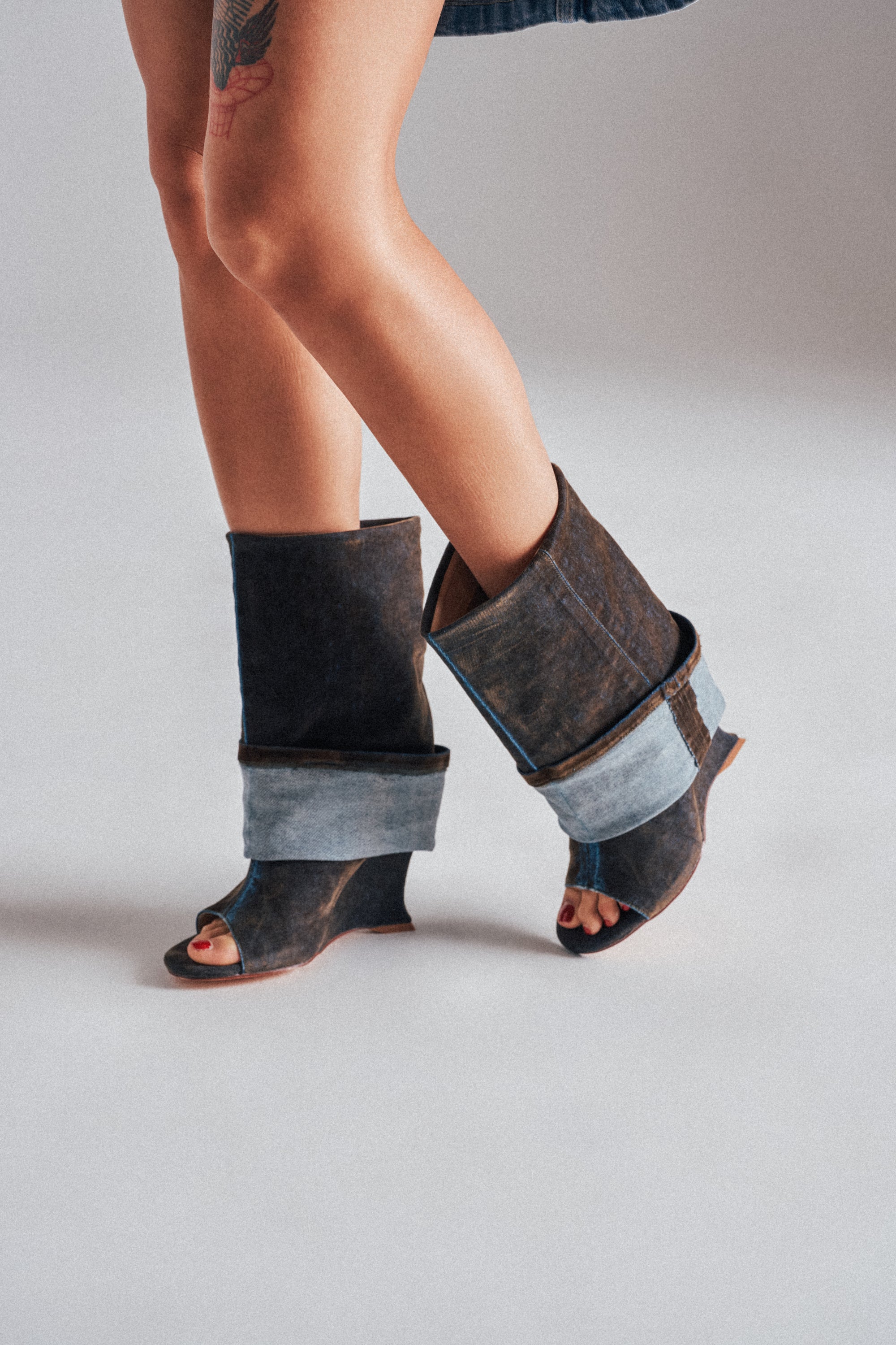 A person wears the AZALEA WANG TIN BROWN CUFFED WEDGE BOOTIE—an open-toe, mid-calf bootie with wide, folded cuffs in denim and brown leather—standing with legs crossed on a light background.