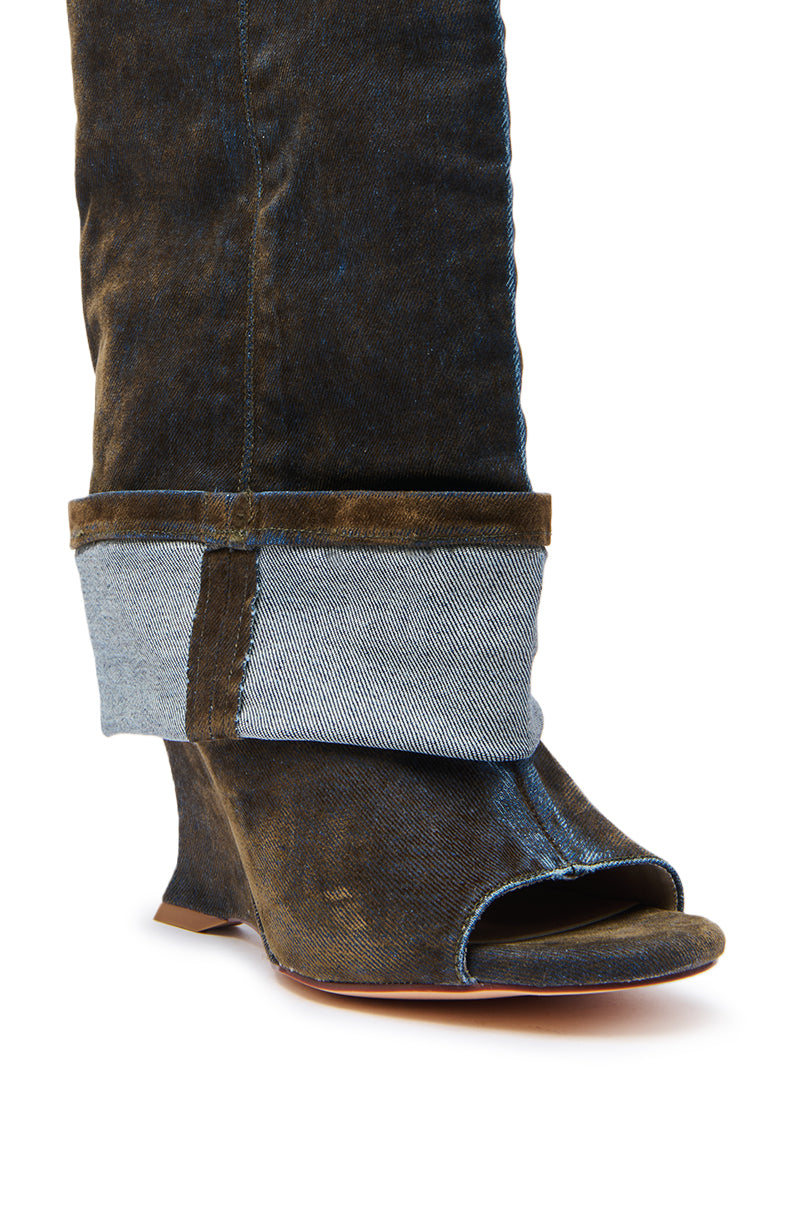 A close-up of a person wearing denim jeans with wide cuffs and the AZALEA WANG TIN BROWN CUFFED WEDGE BOOTIE—a heeled, open-toe wedge rising to mid-calf that perfectly complements the worn texture of both jeans and boots.