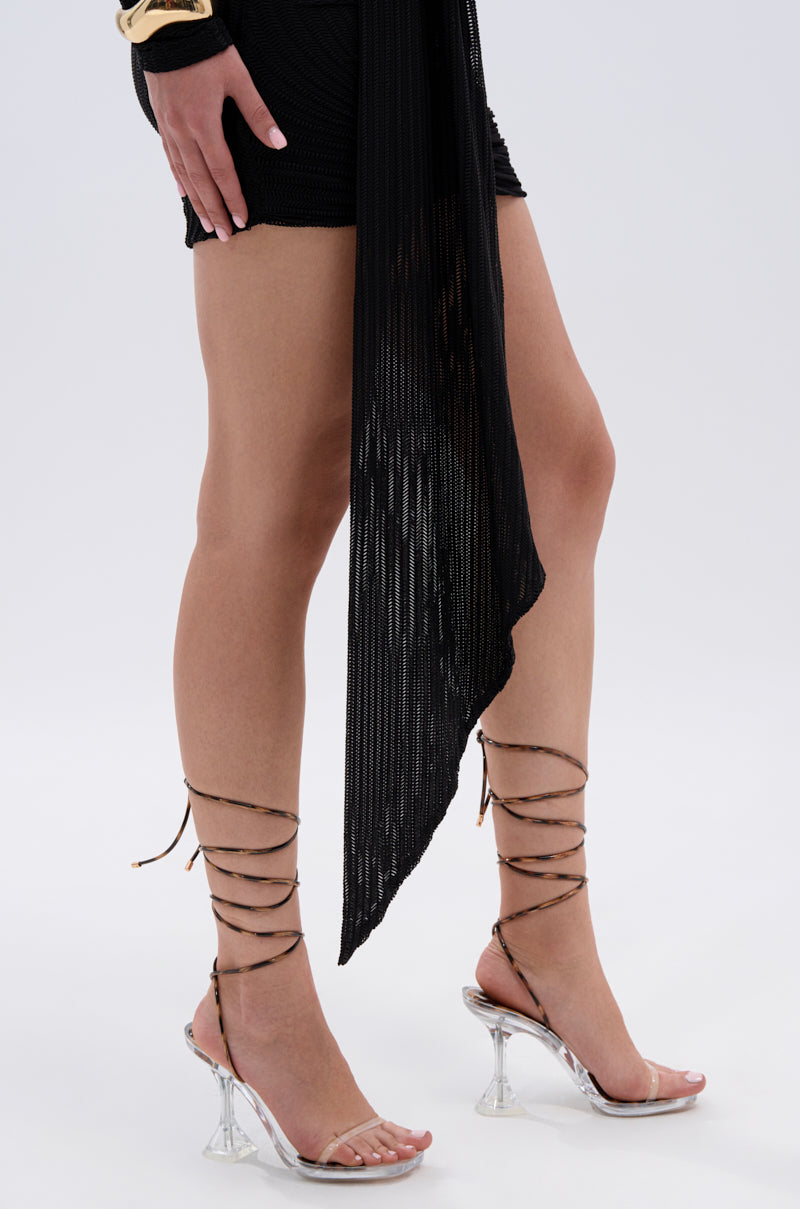 A person wearing a short black skirt, sheer black wrap, and the AKIRA TINKER LEOPARD LACE UP CHUNKY SANDAL stands against a plain light background. Only their lower half is visible, with one hand resting on their hip.