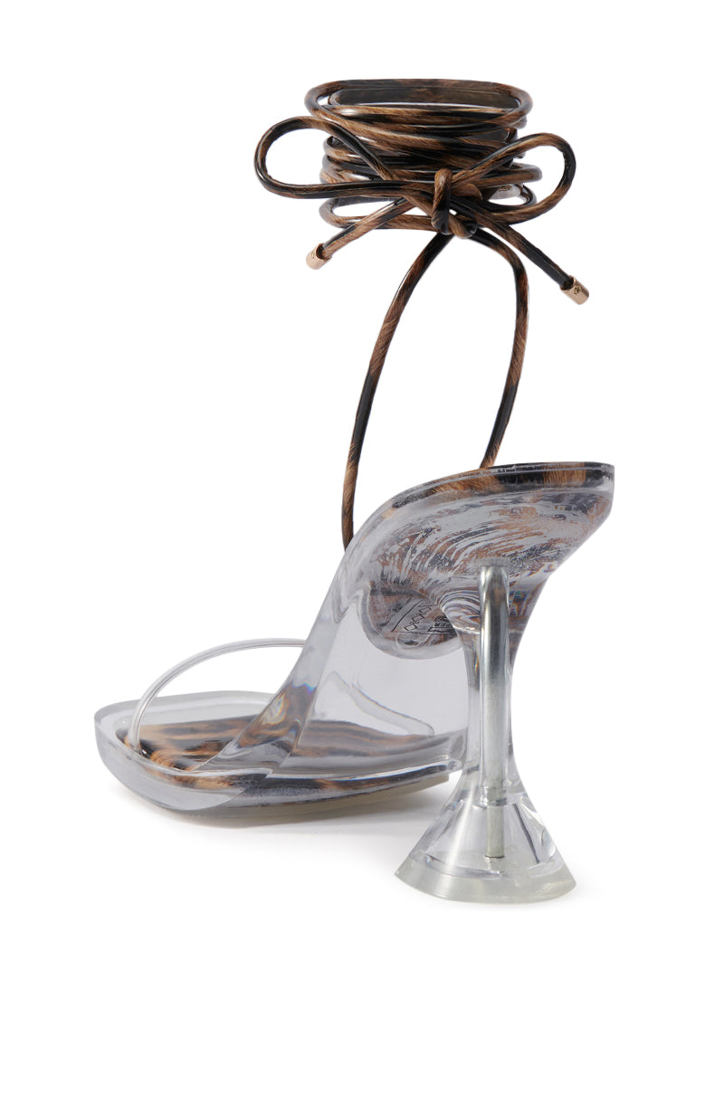 The AKIRA TINKER LEOPARD LACE UP CHUNKY SANDAL features a flared Lucite heel, transparent PVC vamp strap, and brown lace-up straps that wrap around the ankle.
