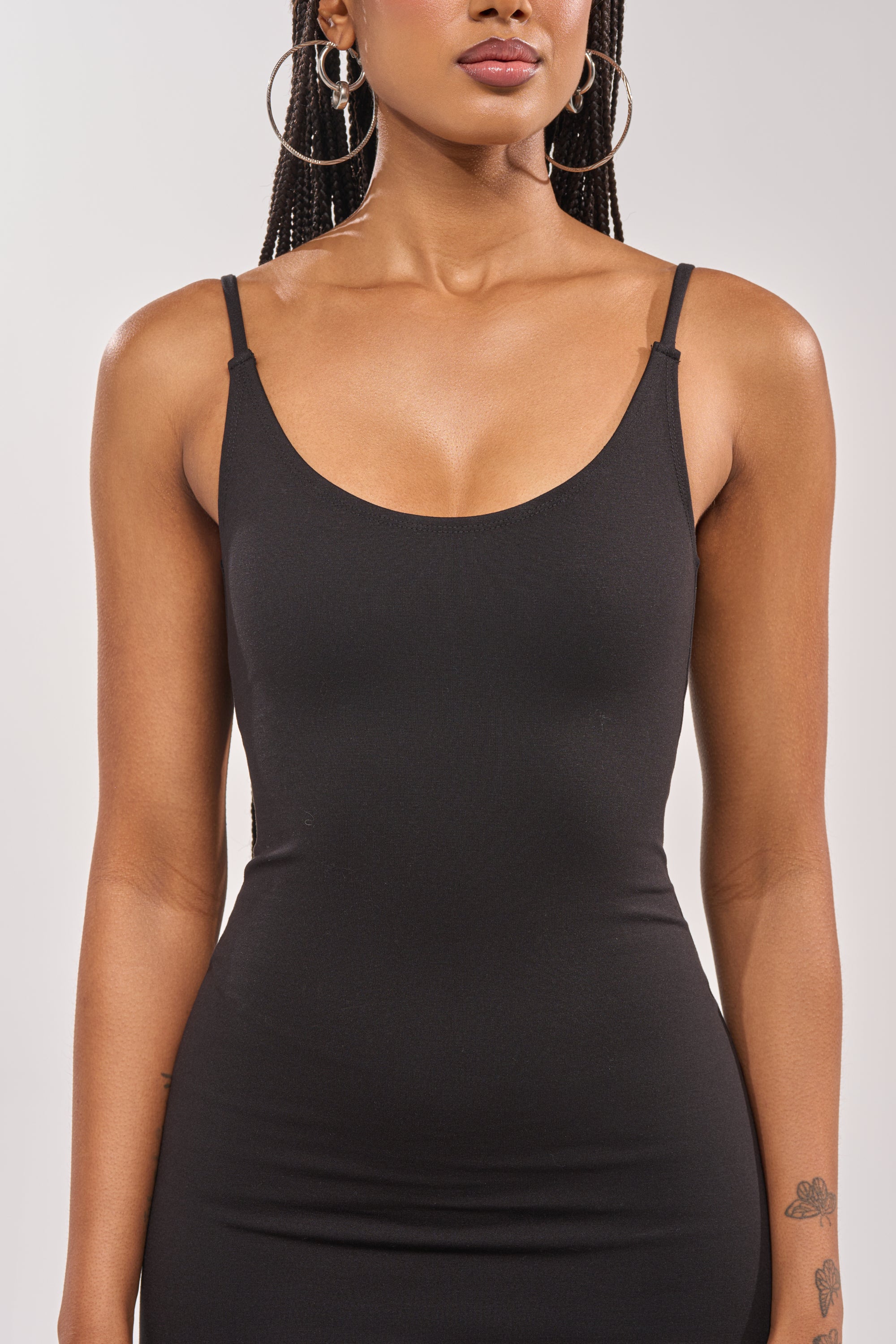 A woman in a sleeveless black dress models the TITSY ADHESIVE BRA, providing a seamless look beneath her outfit, with large hoop earrings and shown from shoulders to mid-thigh against a plain background. 2