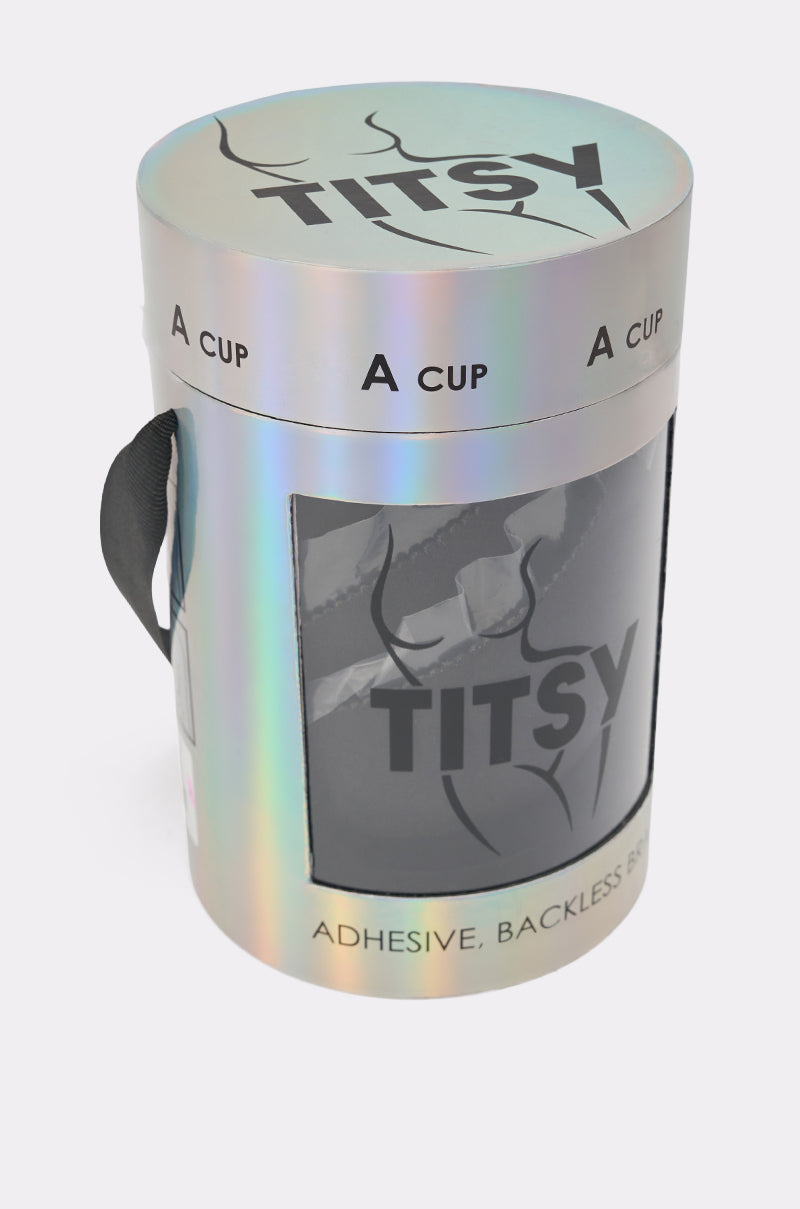 A holographic cylindrical box with a clear window displays the black TITSY ADHESIVE BRA (size A cup). The packaging features a black carrying handle and highlights the seamless, strapless design. 5