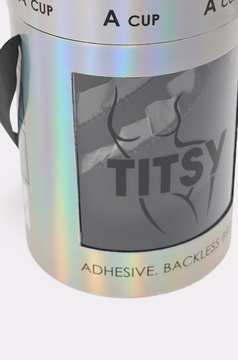 The TITSY ADHESIVE BRA comes in a holographic cylindrical package labeled "A CUP" and "TITSY," featuring a clear window that displays the black, seamless, strapless bra and black line art of a woman’s torso. 6