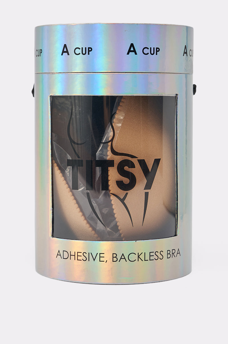 A holographic cylindrical container labeled "TITSY ADHESIVE BRA IN NUDE" displays a nude, adhesive, backless bra for A cups through a clear window, with bold black text and a minimalist bra illustration on the packaging. 5
