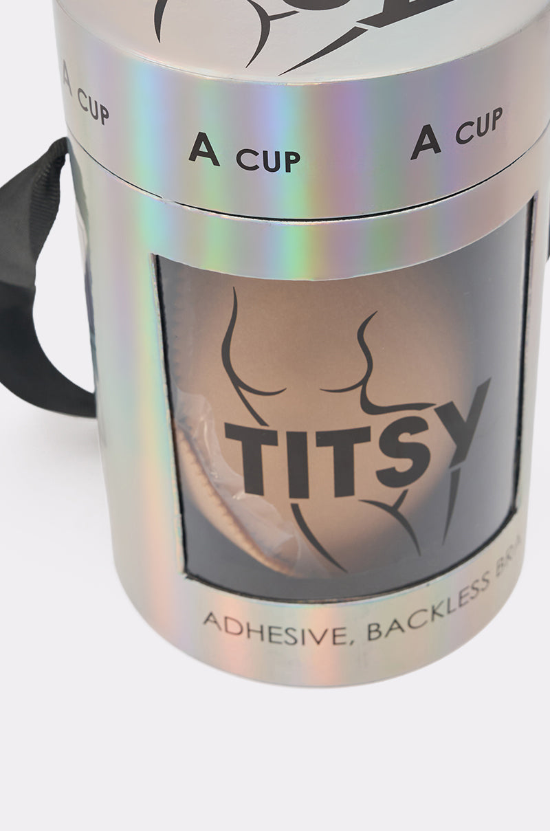 A holographic cylindrical container for TITSY ADHESIVE BRA IN NUDE features “A CUP,” a stylized torso, a black ribbon handle, and details like “ADHESIVE, BACKLESS BO,” hinting at adhesive bra cups. 6
