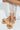 Seated with their feet in TIZZY RAFFIA FLAT BOW SANDALS, a person pairs them elegantly with a white lace skirt. Light pink nails enhance the summer footwear against a smooth, light backdrop.