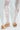 A person in sheer lace pants with intricate floral designs stands on a light surface, complemented by TIZZY RAFFIA FLAT BOW SANDALS. These tan sandals boast a vegan leather insole, offering a casual, elegant summer footwear style.
