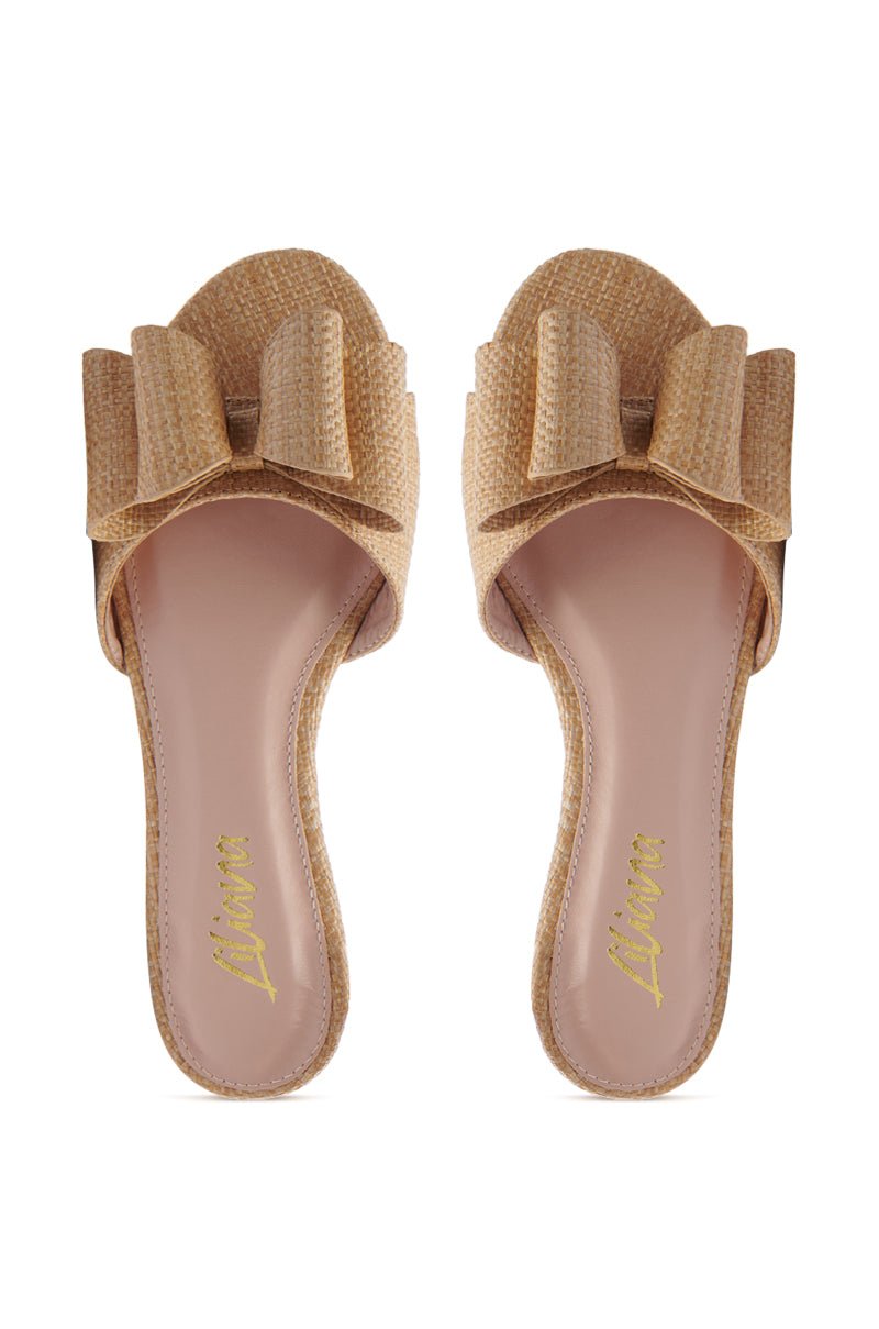 The TIZZY RAFFIA FLAT BOW SANDAL is a beige slip-on with large bows, open toes, and a raffia-inspired woven texture. It features a vegan leather insole with a pinkish tint and golden logo, viewed from above for perfect summer style.