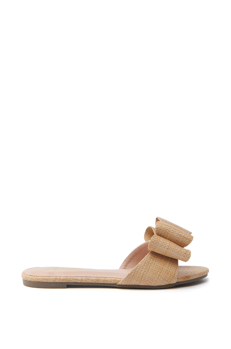 The TIZZY RAFFIA FLAT BOW SANDAL, with its textured surface and large bow on the strap, is ideal for summer. It boasts a tan finish, vegan leather insole, and elegant design on a plain white background.