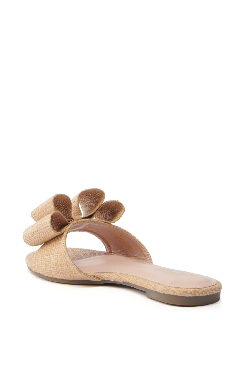 The TIZZY RAFFIA FLAT BOW SANDAL is a tan woven sandal featuring a flat sole, an oversized decorative bow on the upper strap, and a vegan leather insole. Displayed at a slight angle, it's the perfect piece of summer footwear encapsulating effortless style.