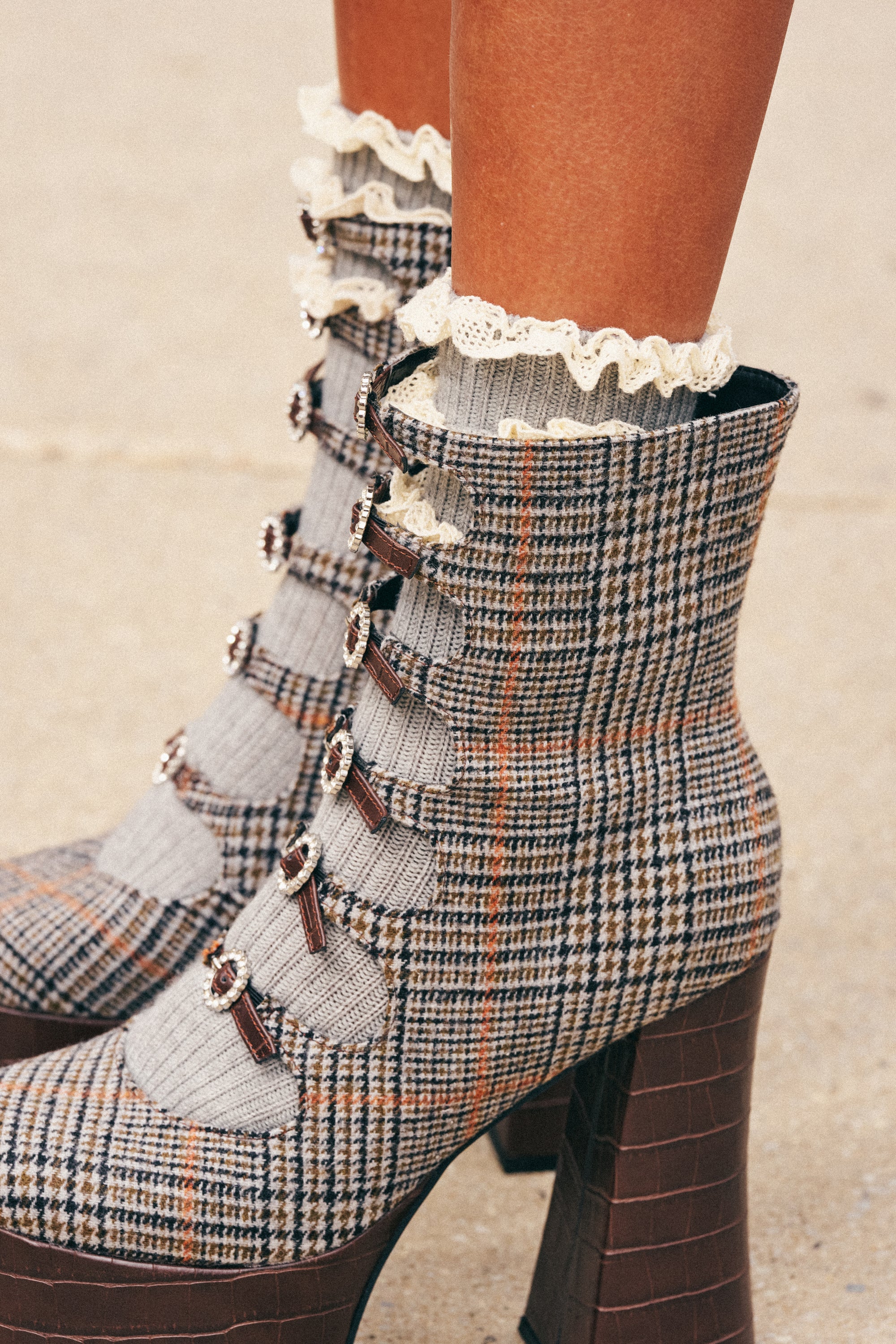 Wearing high-heeled, plaid boots adorned with buckles and decorative buttons, a person stands on a sidewalk with their look completed by the LAYERED CUTIE RUFFLE SOCKS IN GREY, which feature girly lace accents perfect for fall.
