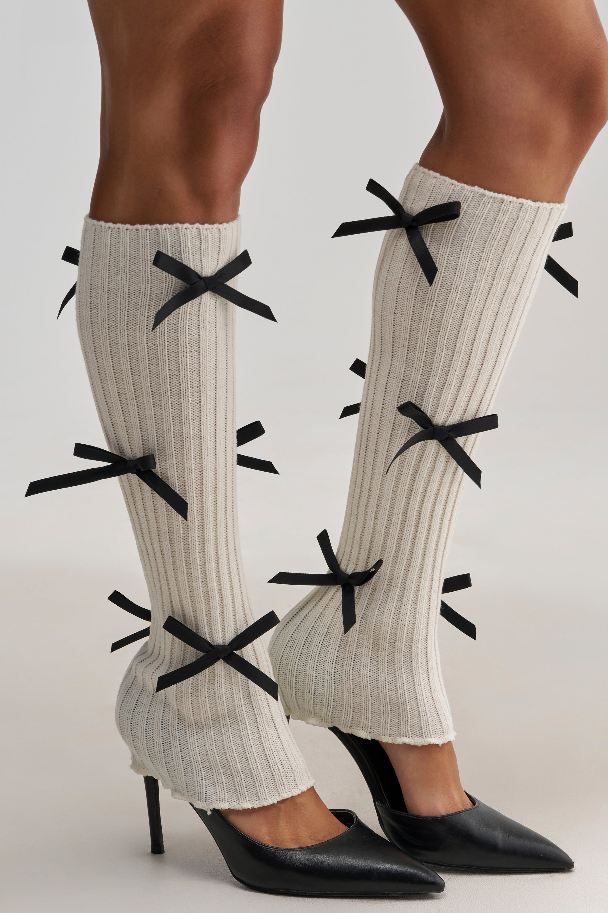A person wears the GIRLY POP LEG WARMERS in beige, featuring black bows, paired with black pointed high heels. The knee-high leg warmers cover the calves and ankles while leaving the heels exposed.