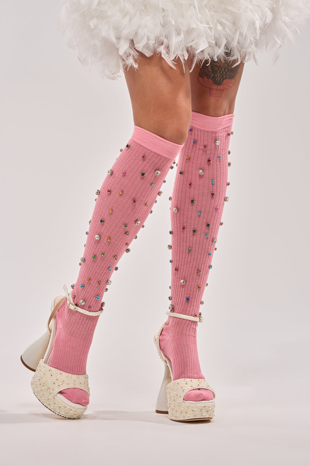 A model stands on a light background wearing COMPLETE THE LOOK EMBELLISHED SOCKS—pink socks adorned with colorful beads—paired with white high-heeled sandals and a white, feathery skirt. 0