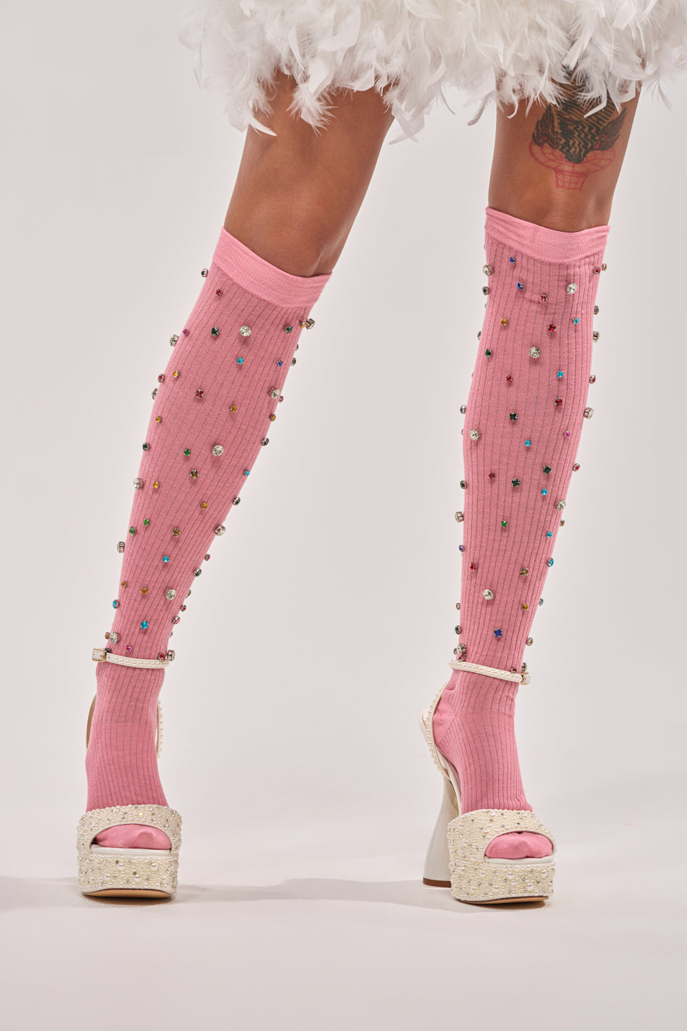 Model wears COMPLETE THE LOOK EMBELLISHED SOCKS—knee-high pink socks with rhinestones—paired with white platform sandals, a white feathered skirt, and shows off a colorful thigh tattoo. 1