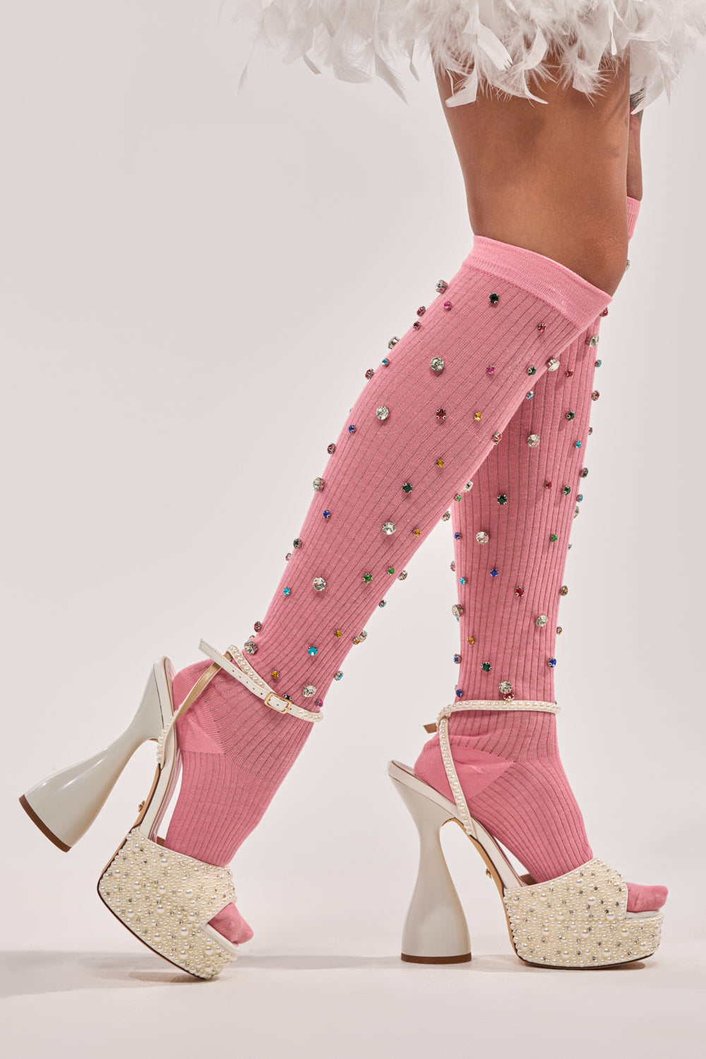 A person models the COMPLETE THE LOOK EMBELLISHED SOCKS—pink, knee-high, and adorned with embellishments—paired with white platform high-heeled sandals and a white feathered skirt hem against a plain background. 2