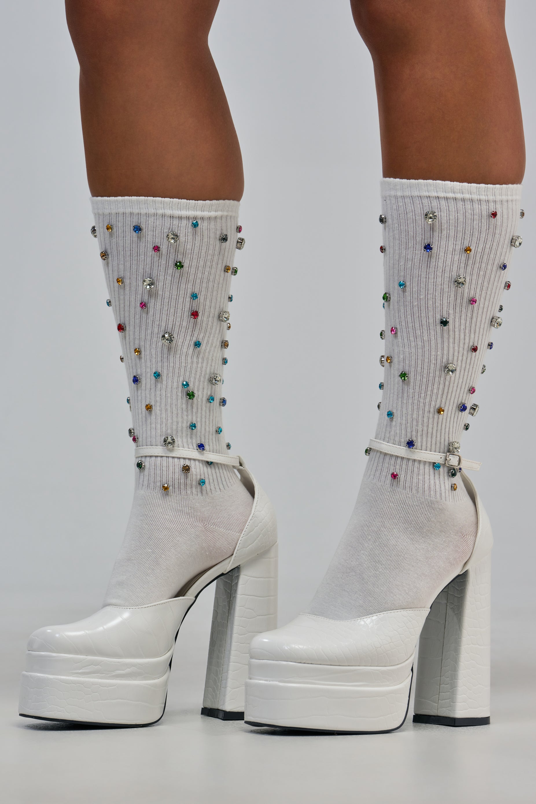 A person stands on a light gray surface wearing white high-heeled platform shoes and the CRYSTAL CONFETTI CREW SOCK IN WHITE—ribbed socks adorned with small, colorful beads.