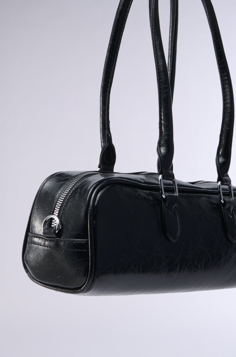 A close-up of the BADDIE BARREL BAG, featuring black faux leather, dual handles, a silver zipper, metal buckles, and a detachable shoulder strap, displayed on a plain light grey background.