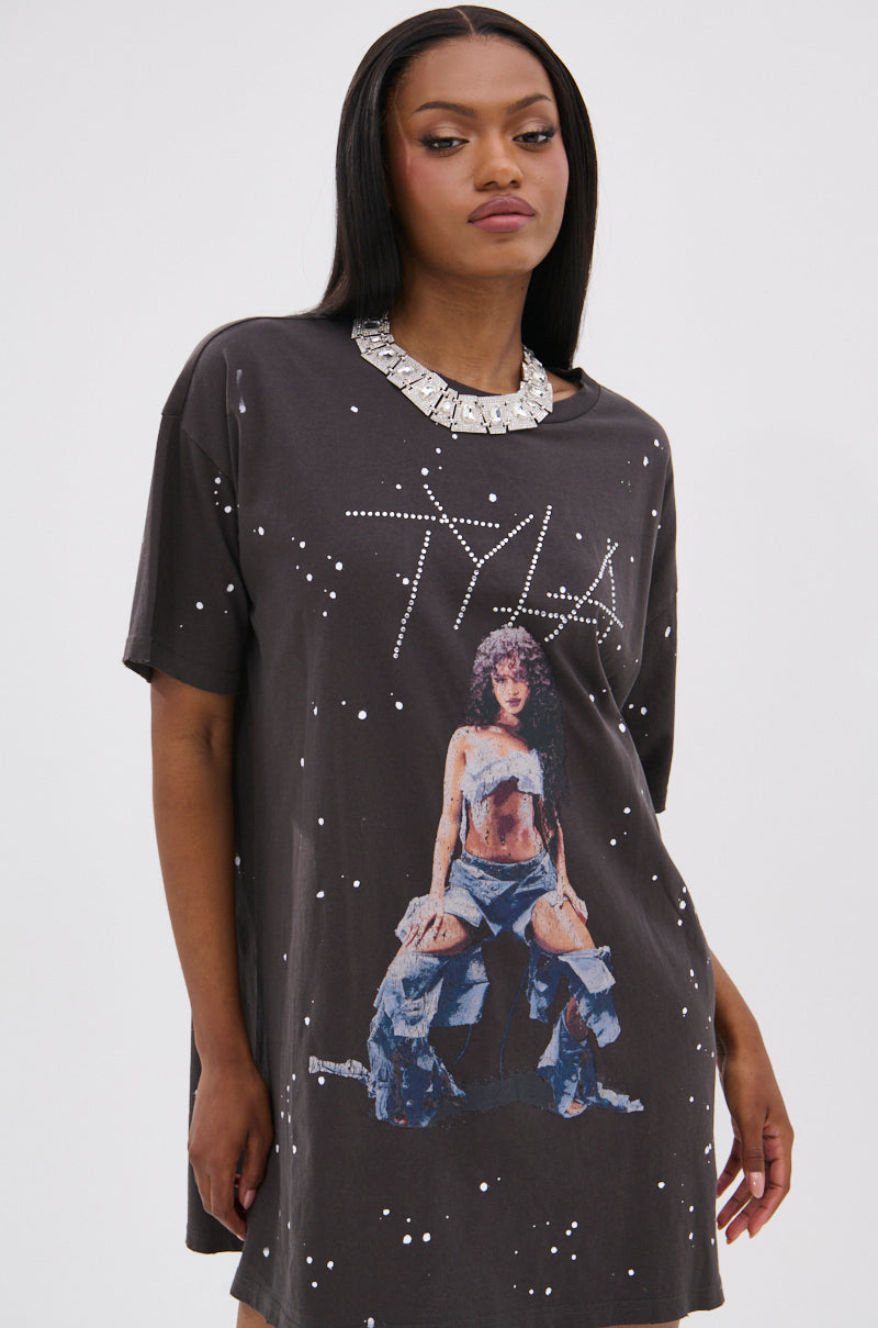 A woman wears the IT GIRL SPLATTERED GRAPHIC TSHIRT DRESS WITH RHINESTONES, featuring bleach splatter cotton, white speckles, a seated woman graphic, and "TYLA" in rhinestones. She pairs it with a chunky silver necklace and has long black hair.