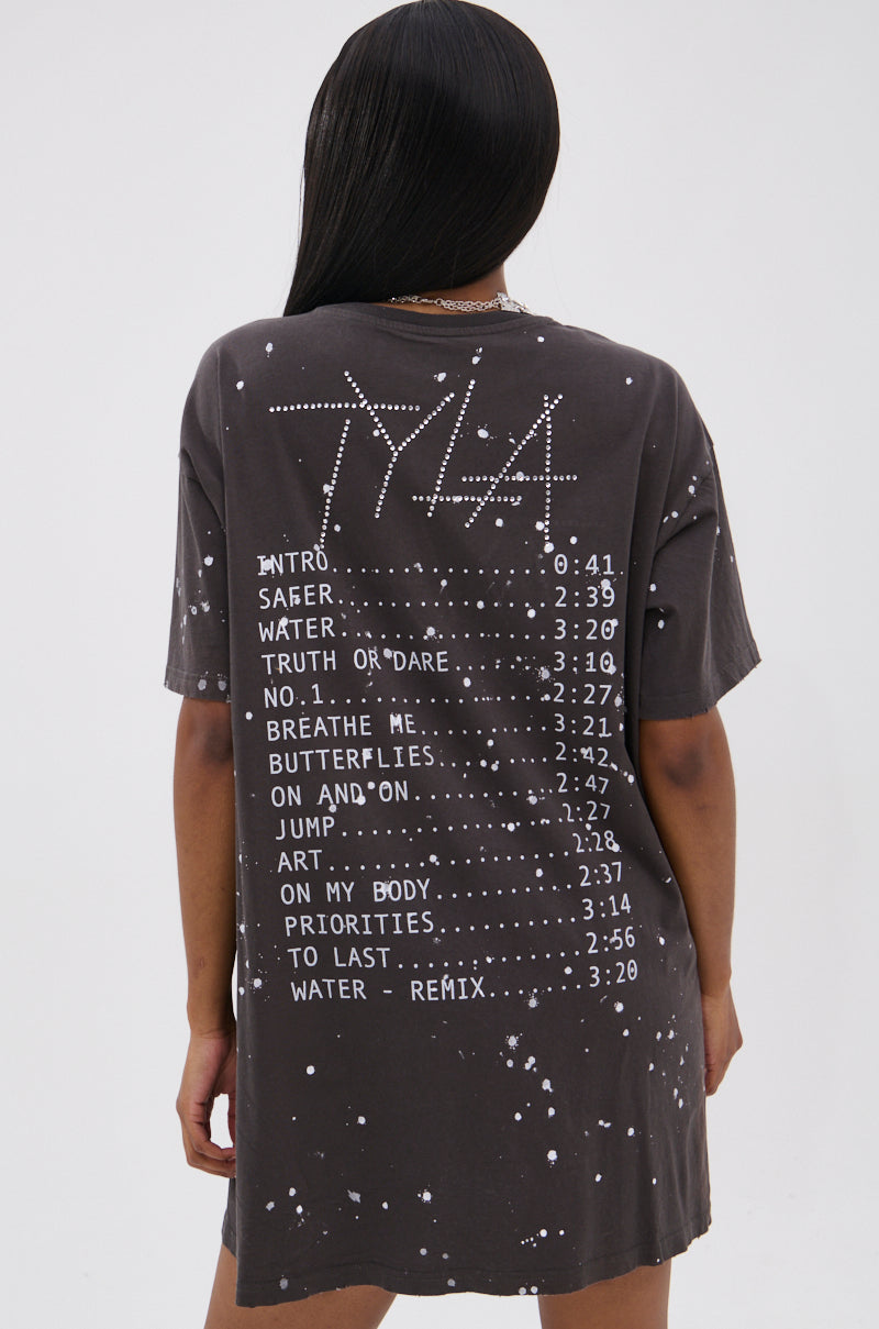 A person with long dark hair faces away from the camera, wearing the IT GIRL Splattered Graphic T-shirt Dress with Rhinestones—an oversized black dress featuring white text, dots, and bold graphic details on the back.