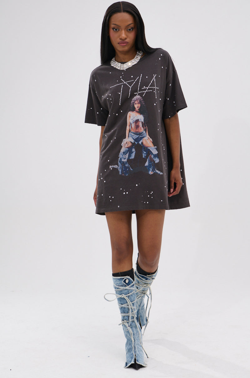 A woman wears the IT GIRL SPLATTERED GRAPHIC TSHIRT DRESS WITH RHINESTONES, featuring an oversized fit, bold graphic print, bleach splatter accents, and rhinestones. She styles it with knee-high lace-up denim boots and a statement necklace.