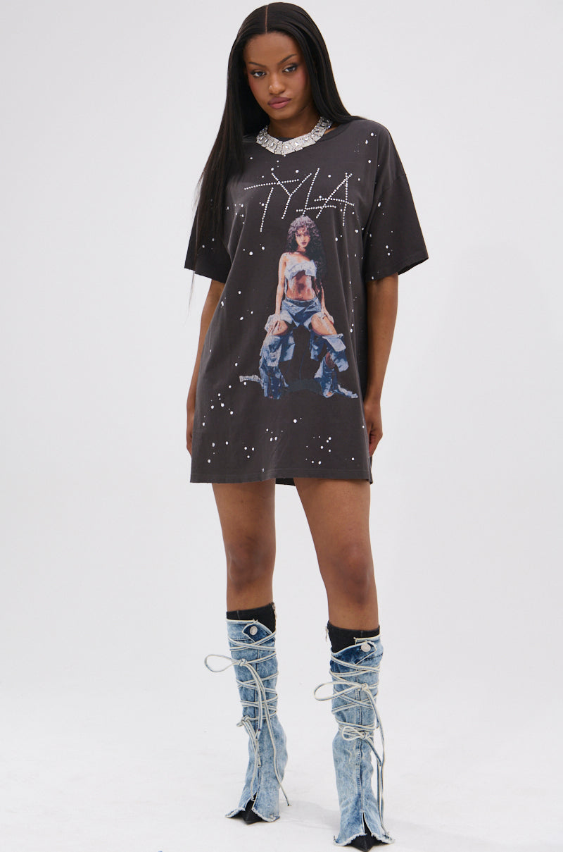 A woman stands against a plain white background wearing the IT GIRL SPLATTERED GRAPHIC TSHIRT DRESS WITH RHINESTONES, accessorized with knee-high light blue denim boots and a chunky silver necklace.