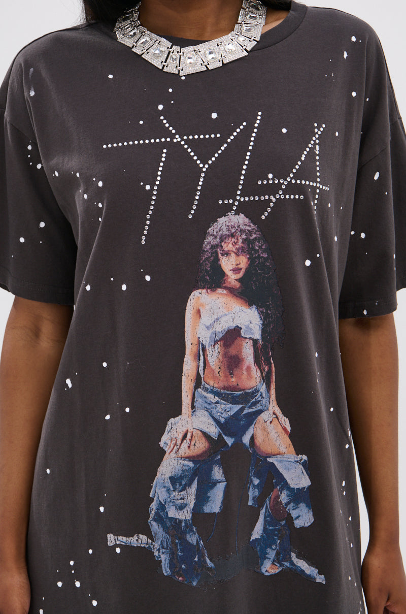 A person models the IT GIRL SPLATTERED GRAPHIC TSHIRT DRESS WITH RHINESTONES, featuring "TYLA" in dotted letters and a bold graphic of a woman with curly hair, silver top, ripped jeans, and sparkling rhinestone accents.
