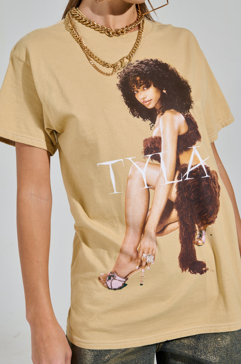 Sporting a beige TYLA GRAPHIC TEE with an image of a woman in a brown furry outfit and "TYLA" boldly printed, accessorized with gold chain necklaces and shiny pants, this look is ideal for fans awaiting Tyla's debut album.