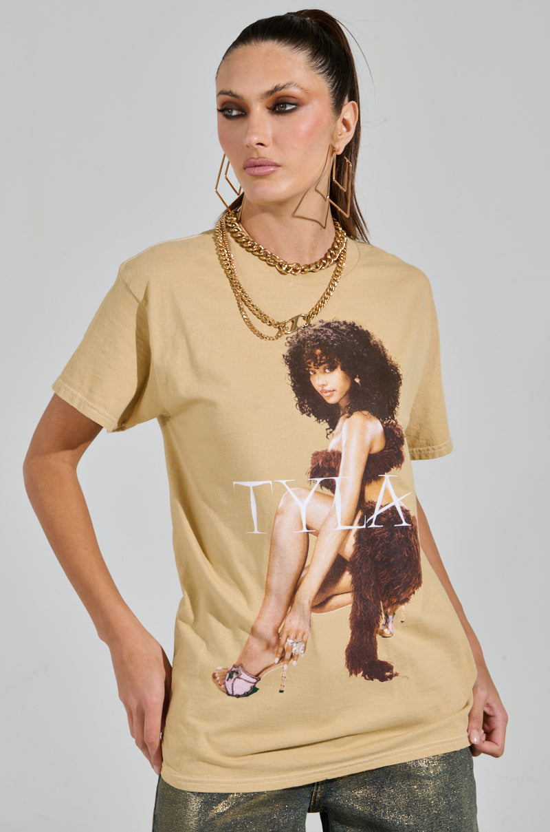 A woman with a high ponytail poses in the beige TYLA GRAPHIC TEE, featuring a curly-haired woman print. She accessorizes with large square earrings and a gold chain necklace, capturing Tyla's vibrant style. One hand rests on her hip.