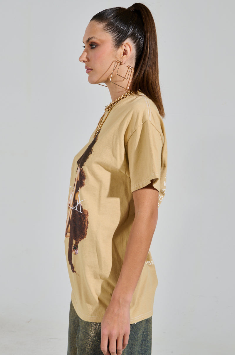 A person with a high ponytail, wearing large geometric earrings and the beige TYLA GRAPHIC TEE with a striking design, stands against a plain background.