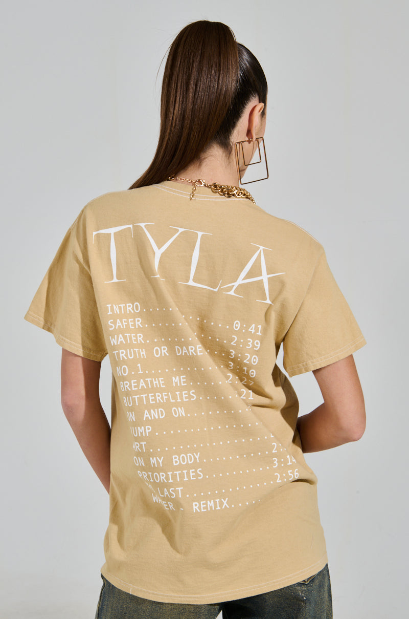 A long-haired person in a ponytail wears a beige TYLA GRAPHIC TEE, featuring the album "Tyla" song titles and times under "TYLA" in bold. They accessorize with gold hoop earrings and a necklace, exuding effortless style.