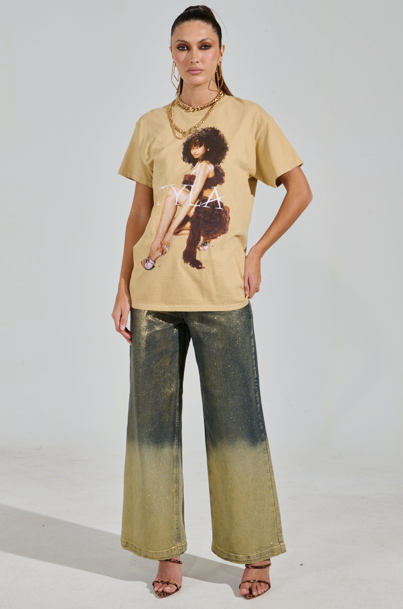 A person stands against a plain background, wearing a Tyla Graphic Tee with an artistic print and wide-leg gradient jeans, accessorizing with gold jewelry and high heels. They pose confidently with one hand on their hip.