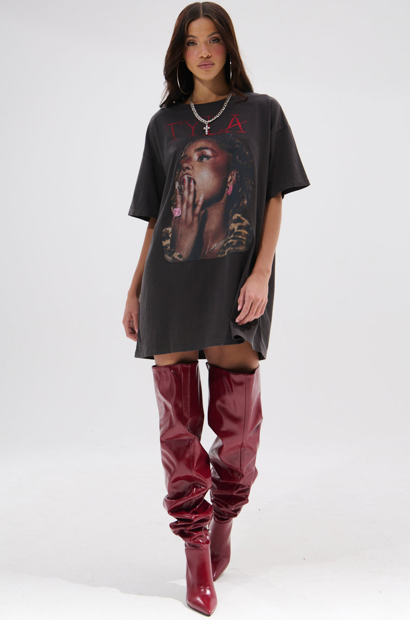 A woman in a TYLA GRAPHIC T-SHIRT DRESS WITH DISTRESSING and shiny, over-the-knee red boots stands confidently against a plain white background.