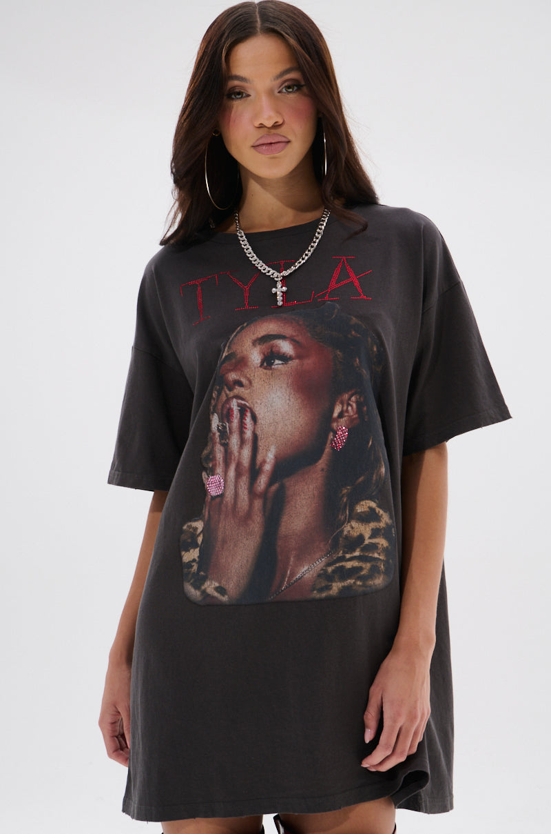 A woman wears the TYLA GRAPHIC T-SHIRT DRESS WITH DISTRESSING, a black dress showcasing a bold woman graphic with pink jewelry, leopard print accessory, and “TYLA” printed in red above the image.