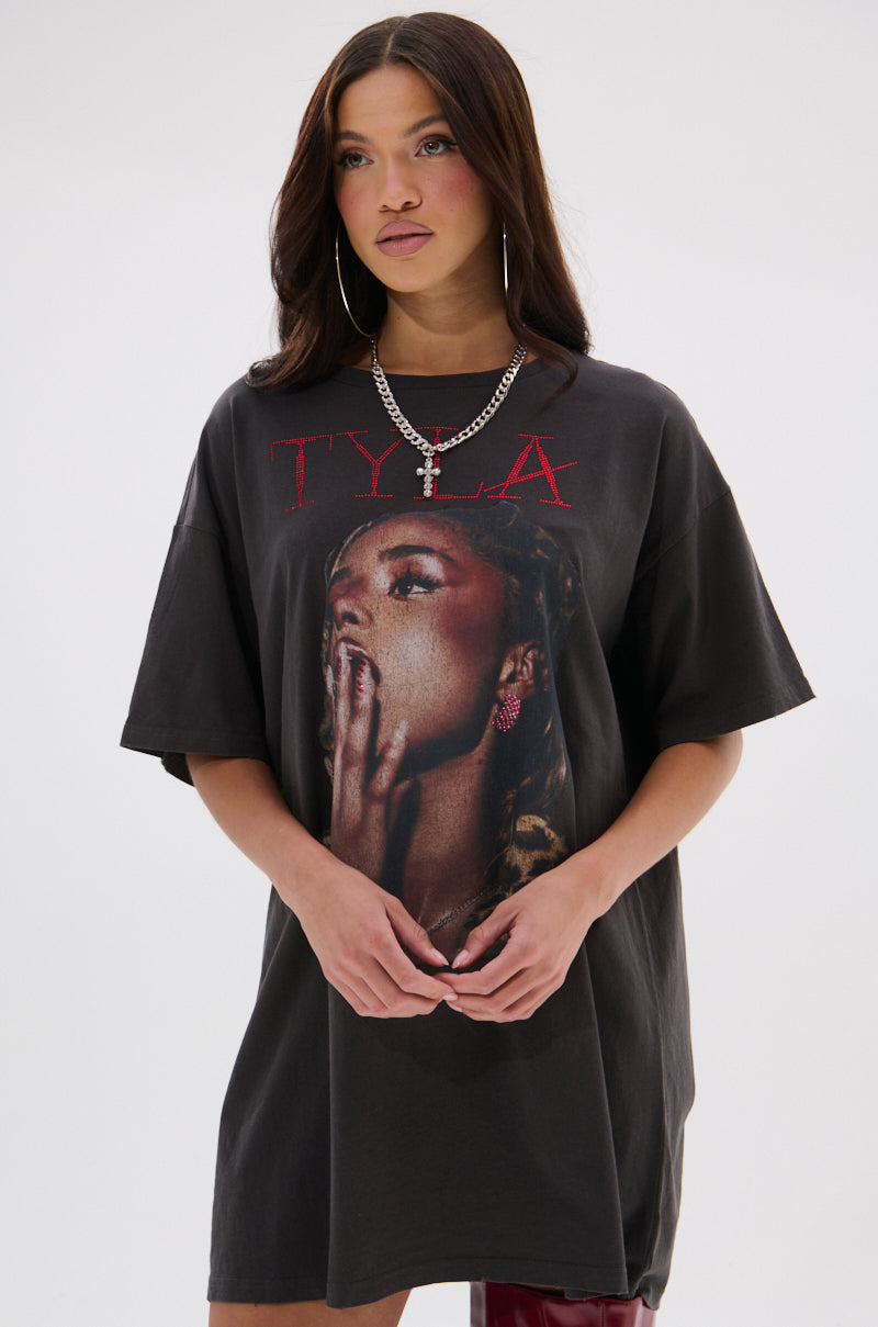 A woman with long brown hair wears the TYLA GRAPHIC T-SHIRT DRESS WITH DISTRESSING, which features a woman's face and red “TYLA” text. She accessorizes with hoop earrings, a chain necklace, and poses against a plain background.