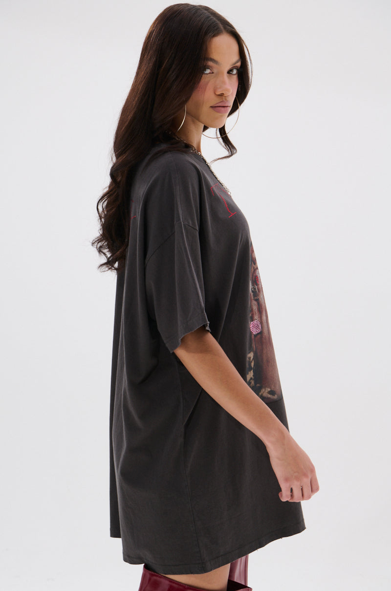 A woman with long dark hair stands sideways against a white background, wearing the TYLA GRAPHIC T-SHIRT DRESS WITH DISTRESSING and red knee-high boots, looking over her shoulder at the camera.