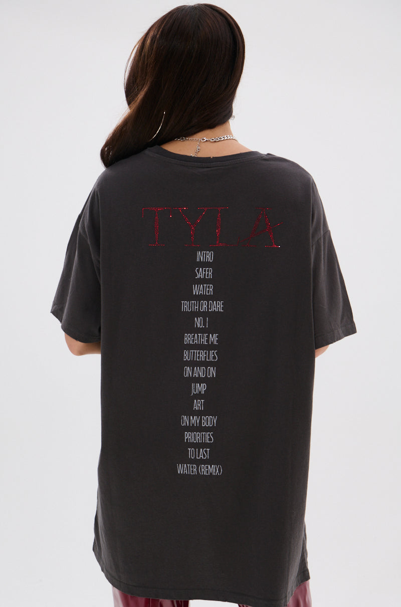 A person with long dark hair, seen from behind, wears the TYLA Graphic T-Shirt Dress with Distressing, featuring "TYLA" in red and white song titles on the back. The background is plain and light-colored.