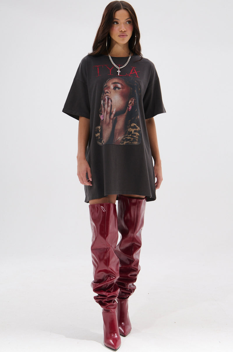 A woman with long, dark hair wears the TYLA GRAPHIC T-SHIRT DRESS WITH DISTRESSING as a dress, paired with shiny thigh-high red boots and a necklace, standing against a plain background.