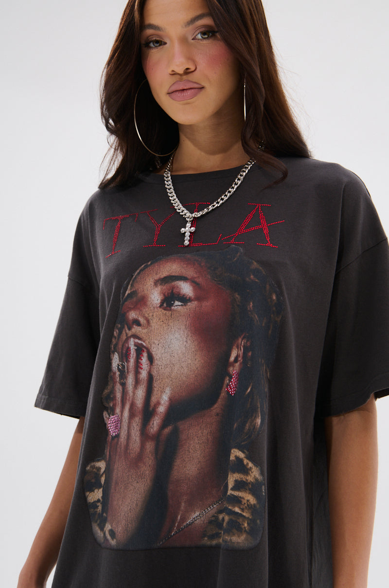 A woman with long brown hair and hoop earrings wears the TYLA GRAPHIC T-SHIRT DRESS WITH DISTRESSING, featuring a thoughtful woman’s pose and bold red "TYLA" lettering above the image.