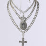 The MADONNA RHINESTONE NECKLACE SET IN SILVER showcases a stunning array of layered silver chains adorned with a decorative cross and a medallion featuring an intricate figure. These curb chain necklaces differ in design and thickness, forming an overlapping, elaborate display against a white background, enriched by religious charm accents.