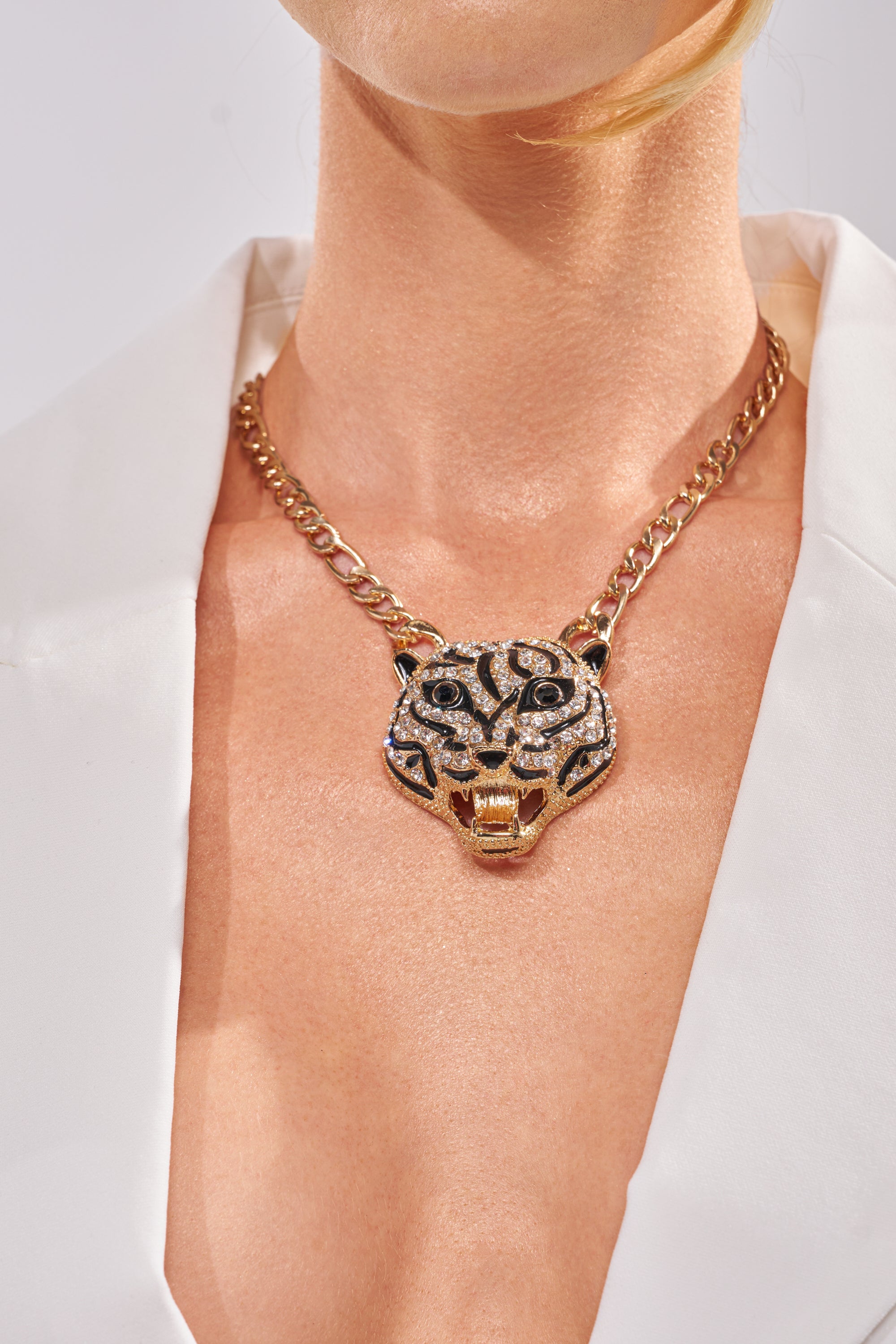 A person wears the HEAR ME NECKLACE—a striking rhinestone tiger piece—layered with a gold chain and styled with a white deep V-neck blazer, drawing all attention to the dazzling necklaces and neckline. 1