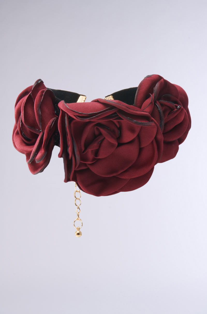 Set against a plain gray background, the IN BLOOM CHOKER NECKLACE features deep red fabric roses on velvet with a delicate gold chain hanging from the center.