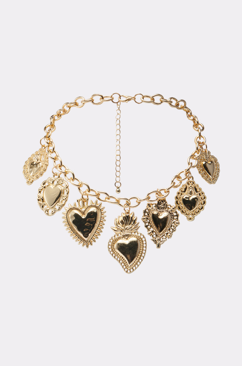 The RAE CHARM NECKLACE is a gold chain with seven ornate heart charms, each with unique vintage-inspired designs, displayed against a white background—an ideal statement jewelry piece.