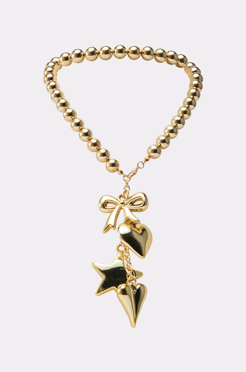 The COQUETTE CHARM NECKLACE is a gold beaded choker with a bow charm, plus dangling star and two heart charms in shiny gold metal, shown against a plain white background.