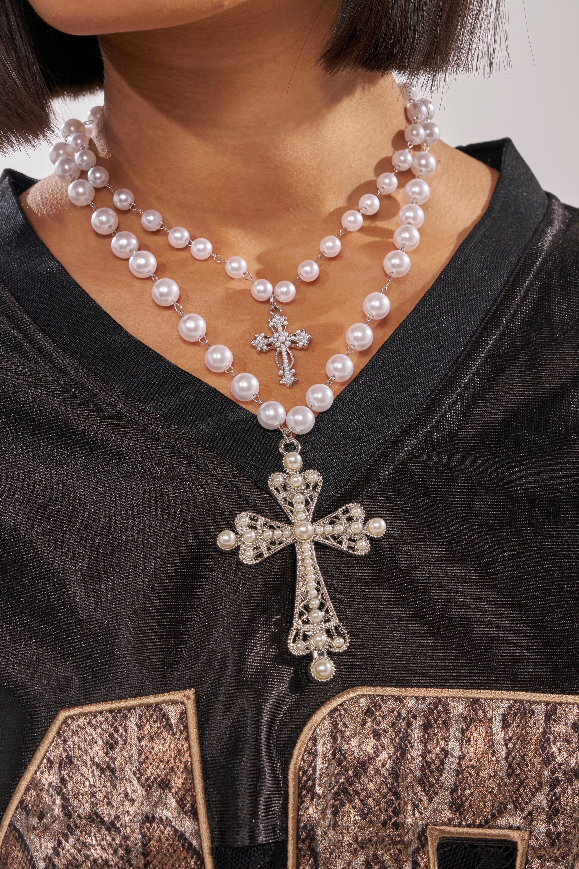 A person wears the IN MY DREAMS NECKLACE—layered strands with cross pendants, one large, one small—over a black shirt with gold numbers. The face isn’t visible. 1