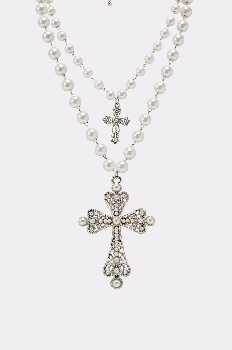 The IN MY DREAMS NECKLACE features two layered faux pearl strands with silver, gem-studded pearl cross pendants—the larger cross hangs lower and the smaller sits above—both elegantly displayed on a plain white background. 3
