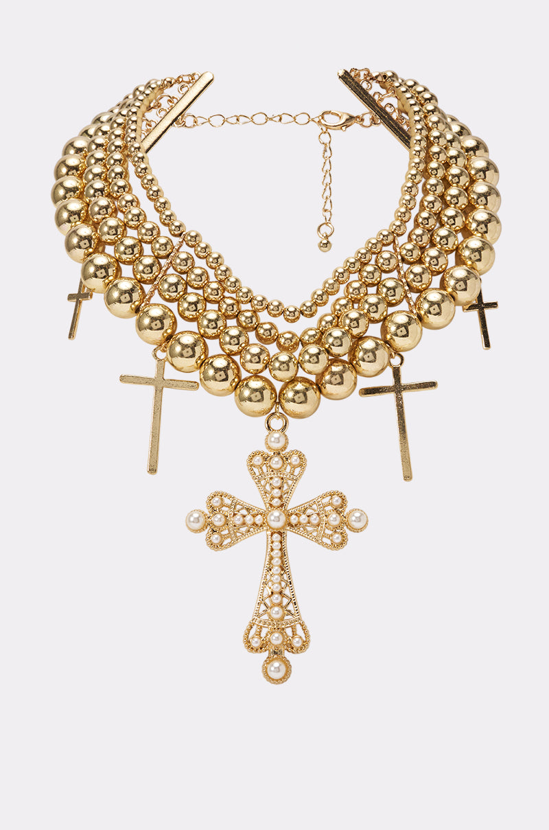 The FESTIVAL DRIP NECKLACE features multiple rows of gold beads adorned with five cross pendants—one large and four smaller—each embellished with pearl-like accents. Set against a white background, this eye-catching necklace is the perfect statement concert jewelry, delivering a bold yet elegant twist on the cross charm necklace trend.