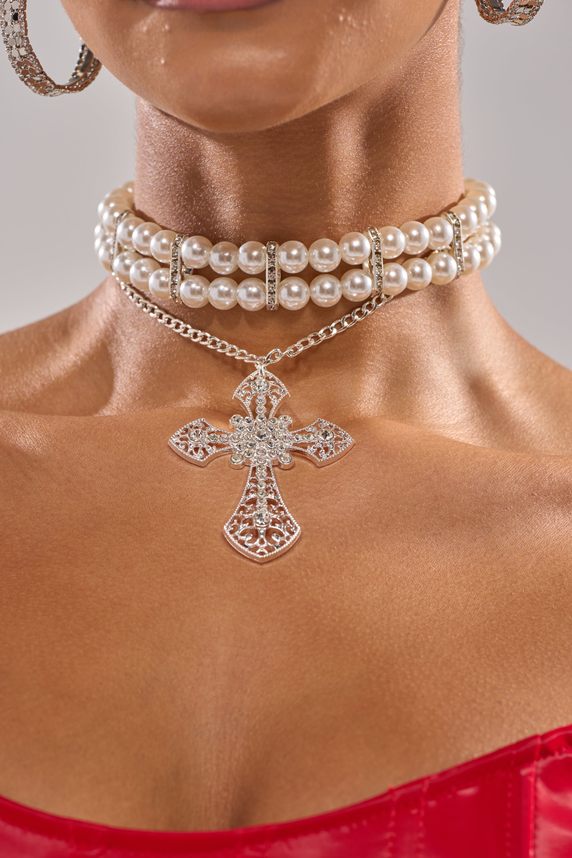 A close-up of a woman’s neck wearing the PURE CROSS NECKLACE, a pearl choker with rhinestones, large hoop earrings, and a red strapless top. 1