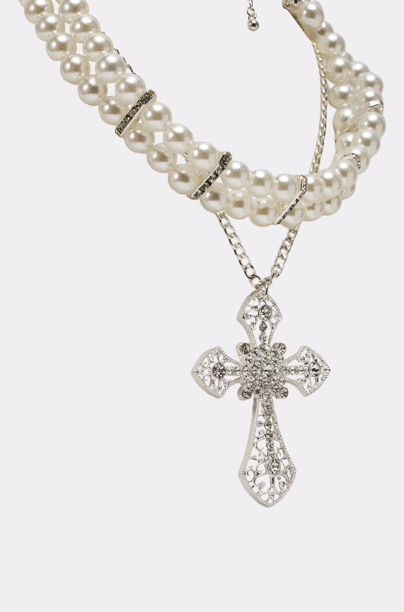 The PURE CROSS NECKLACE features three strands of white pearls with silver spacers and a large ornate silver rhinestone cross pendant adorned with sparkling stones, set against a plain white background. 3