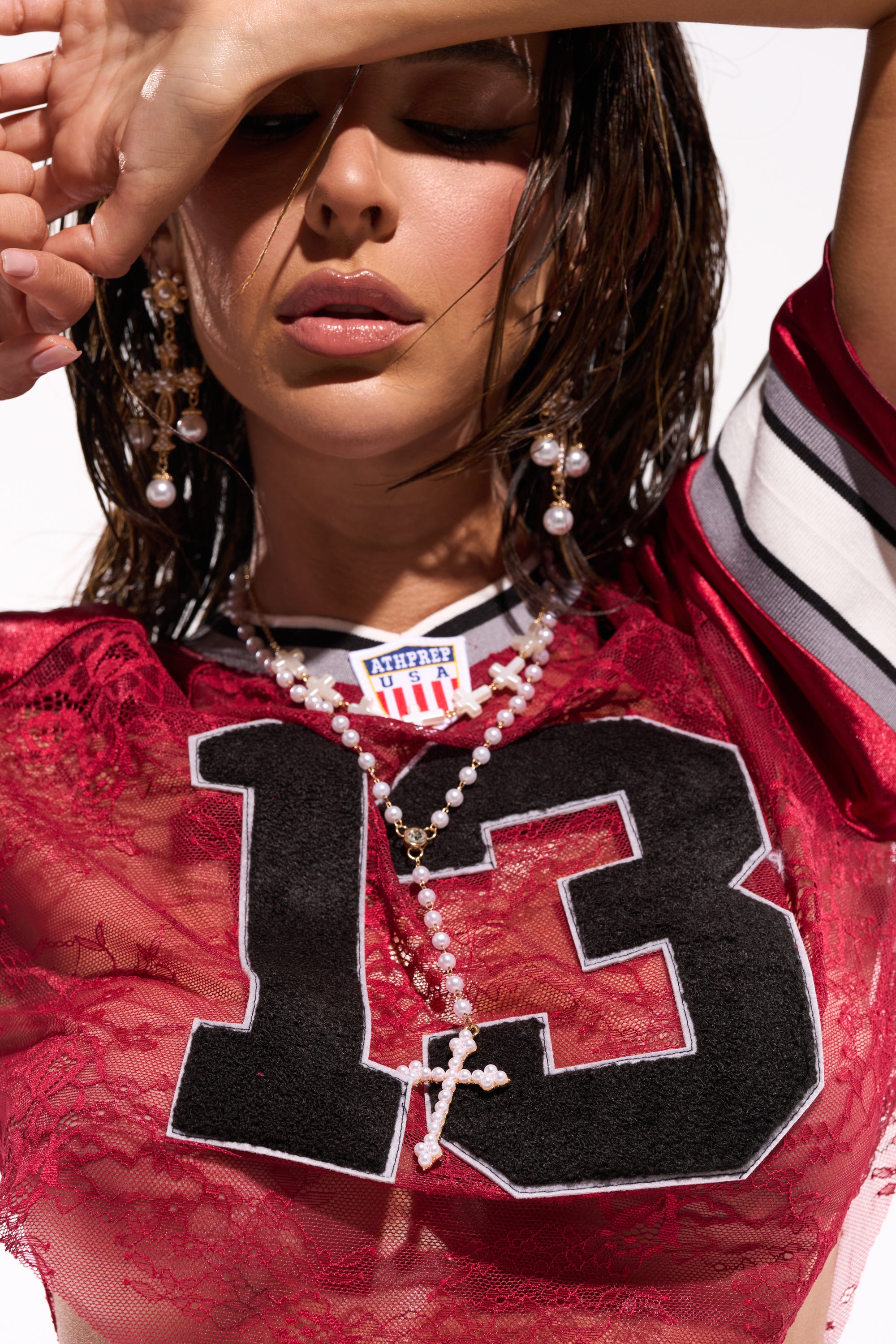 A woman wearing a red sheer top with "13" on it accessorizes with the IN YOUR DREAMS NECKLACE and matching earrings, raising her arm to partially shield her face from the light. 0